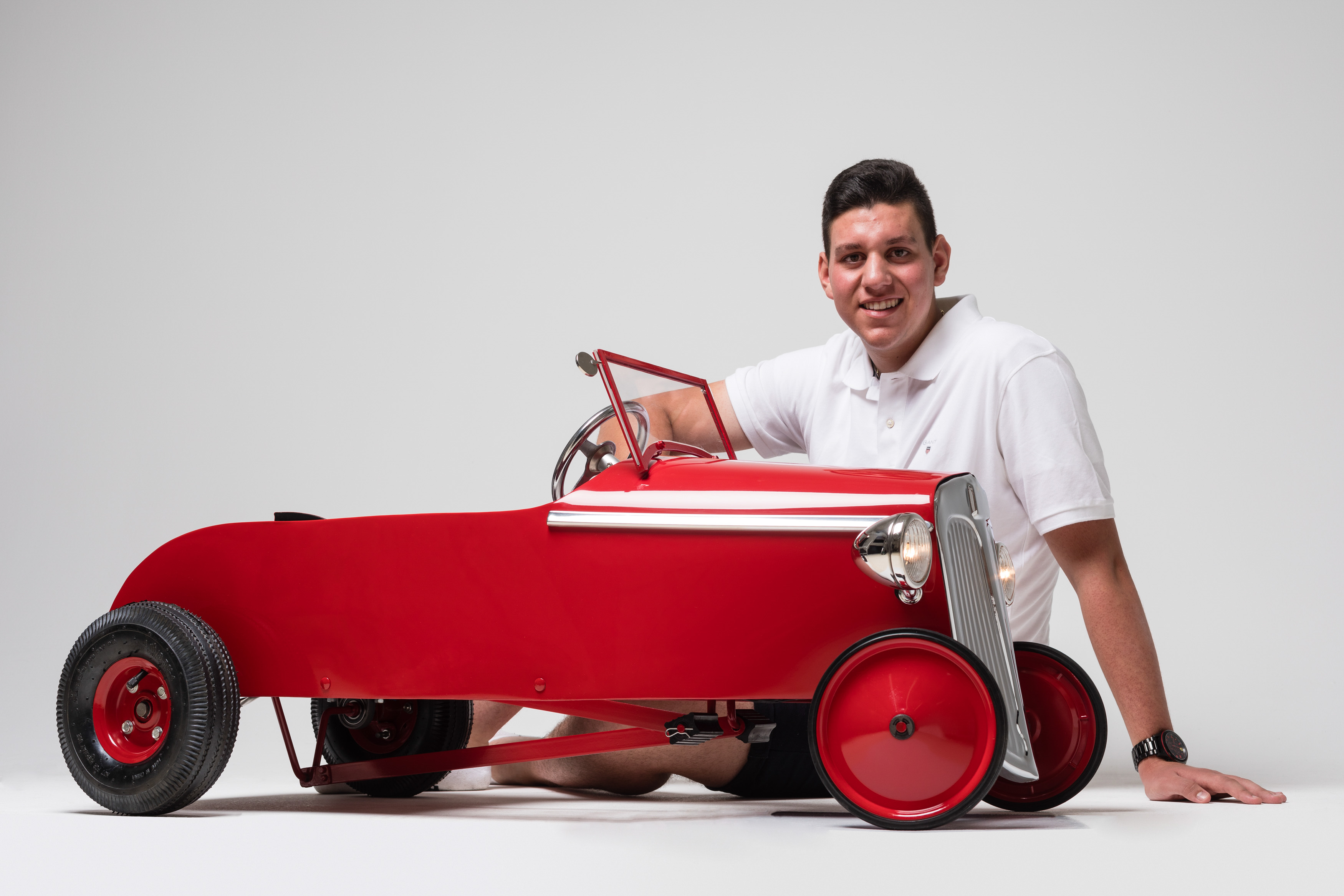 A young man sitting on the floor behind a small, bright red pedal car.